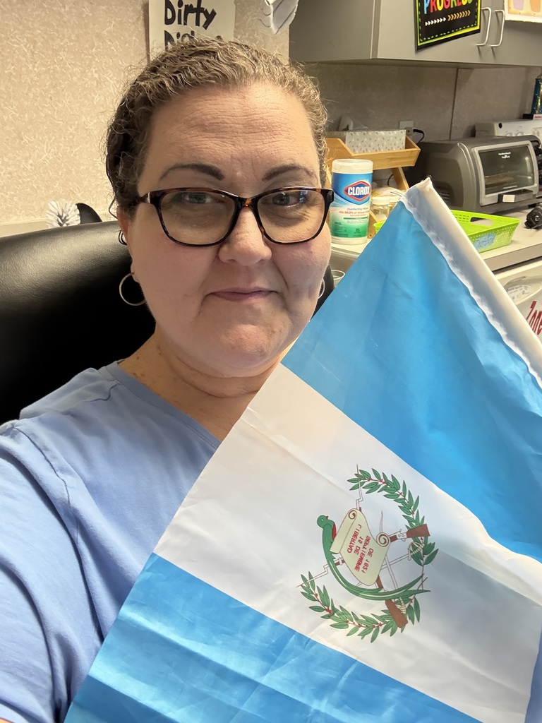 A woman in a blue shirt and glasses holds a blue and white flag with a crest.