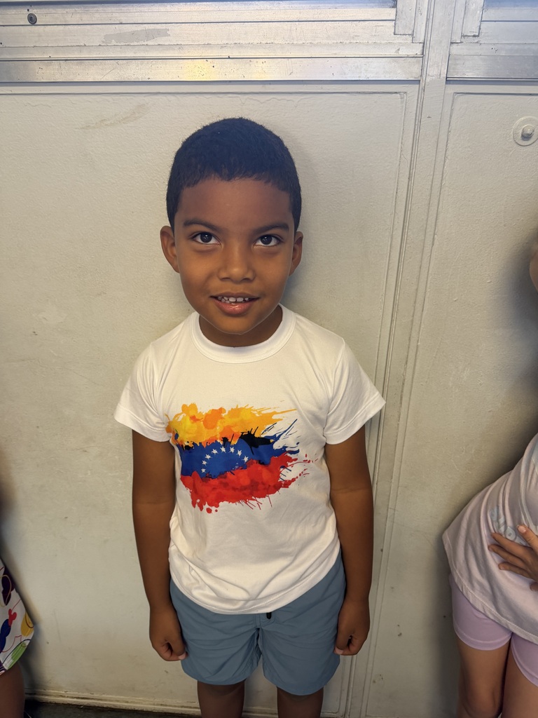 A young boy in a white t-shirt with a colorful design stands against a white wall.