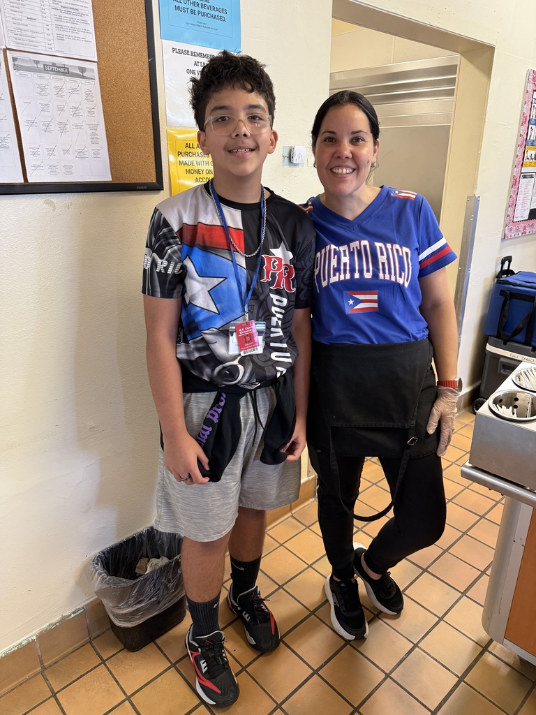 Two individuals stand in a hallway. A boy wears a Puerto Rican flag tee and gray shorts. A woman wears a blue jersey and black pants. A trash can is on the floor.