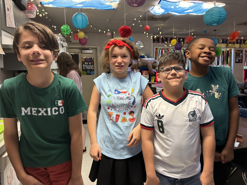 Four young students in a brightly decorated room with balloons and lights. Two wear glasses, one wears a headband, and one wears a green Mexico shirt.
