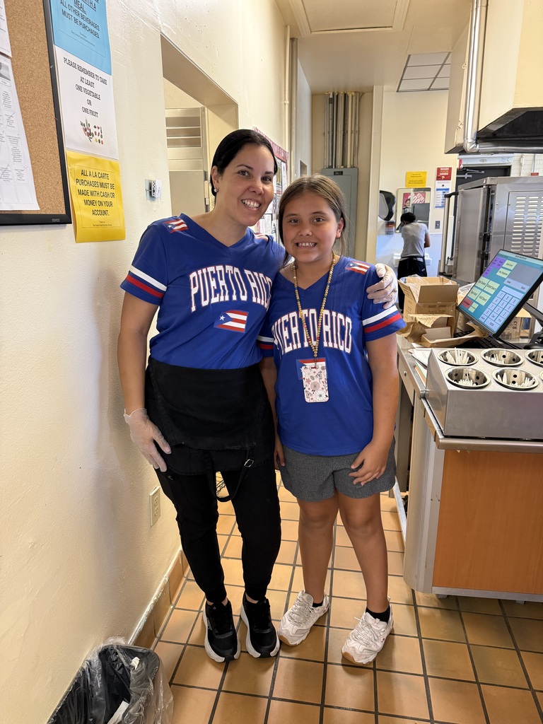 Two women in blue jerseys, one holding a young girl, stand in a hallway with tiled floors and a kitchen behind them.
