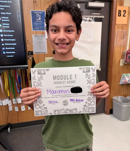 A boy with a smile holds a white certificate with the words "Module 1 Highest Score" in a classroom.