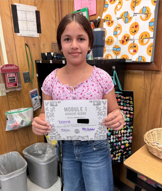 A young girl holds a certificate in a classroom. She wears a pink top, jeans, and a necklace. Behind her, various items are on shelves and a desk.