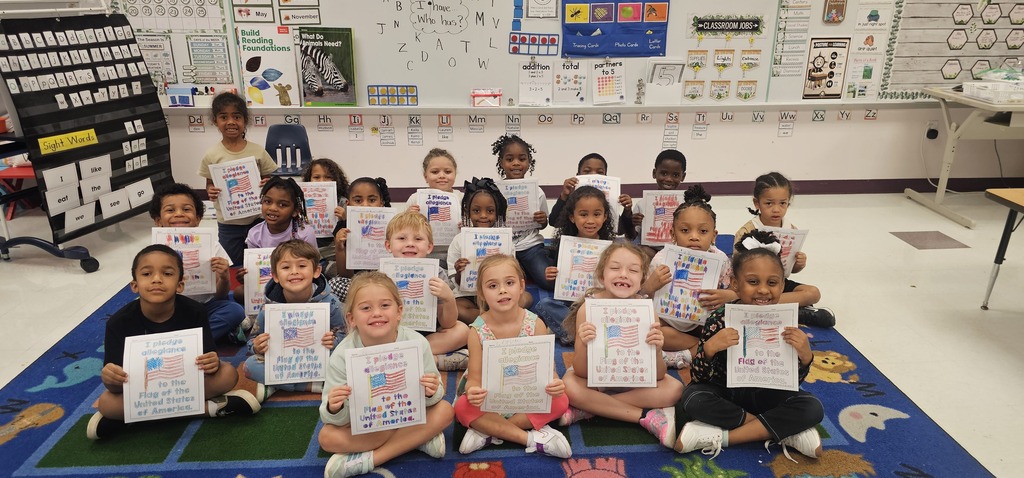 Group of children in a classroom hold up white papers with drawings and writing. A blue rug covers the floor.