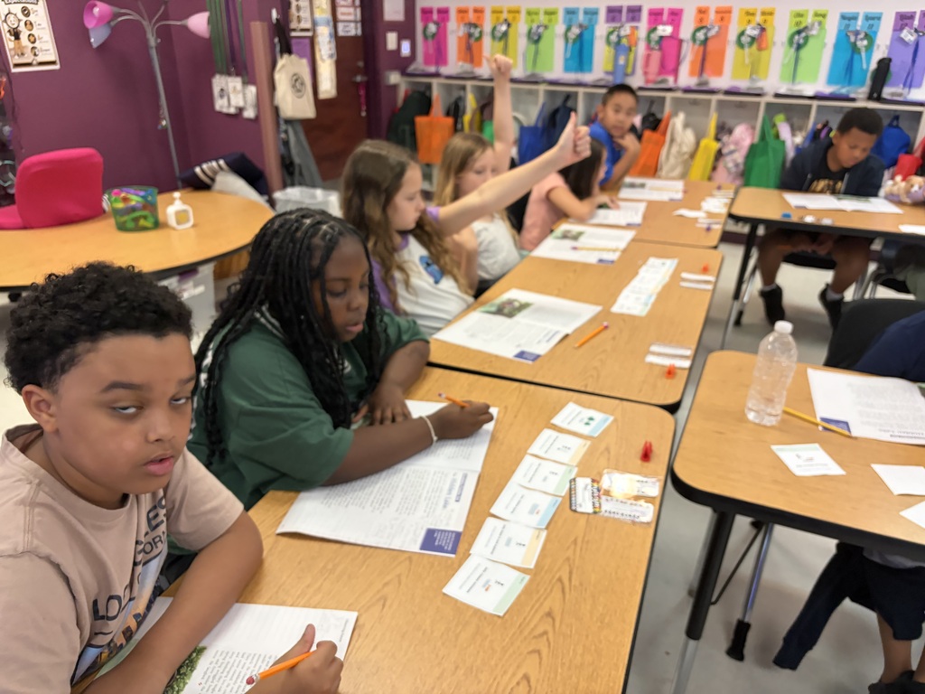 Kids at desks, engaged in an activity with papers and pens. The classroom has colorful charts on the walls.