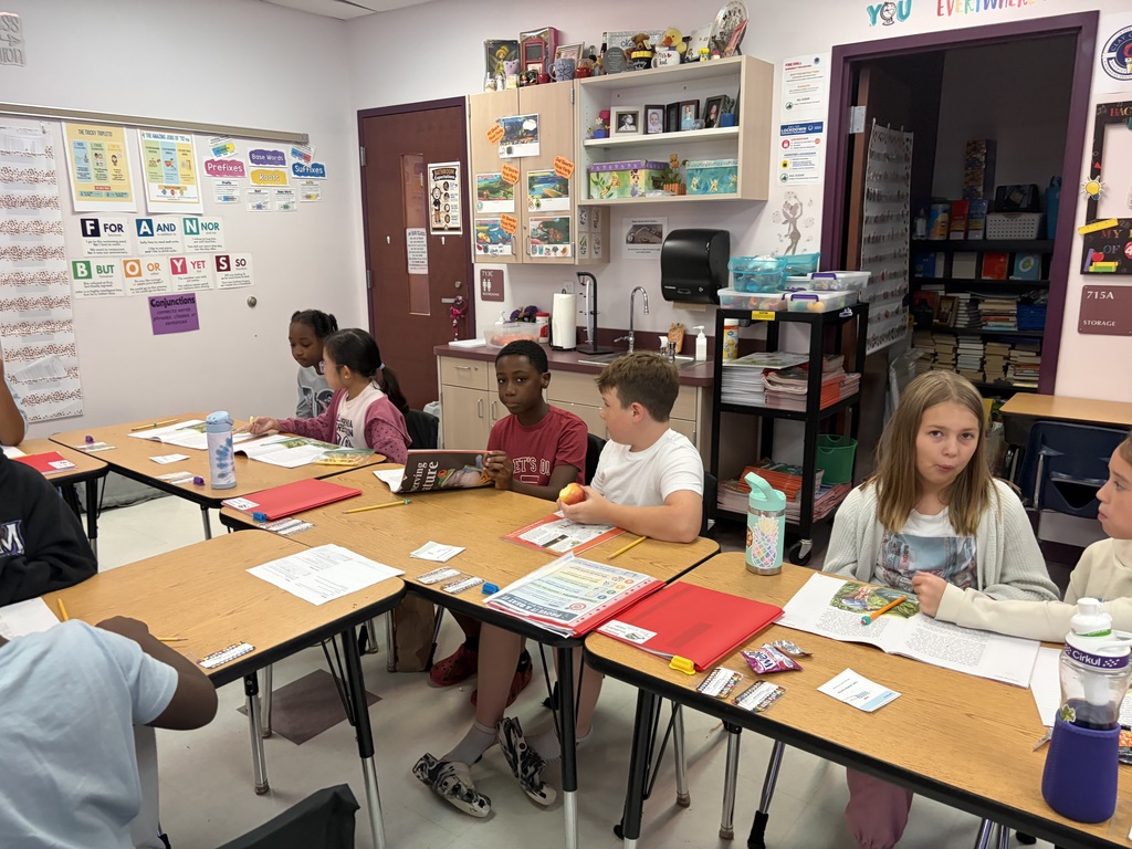 Kids at tables in a classroom, working on projects and reading. Some have books, papers, and water bottles.