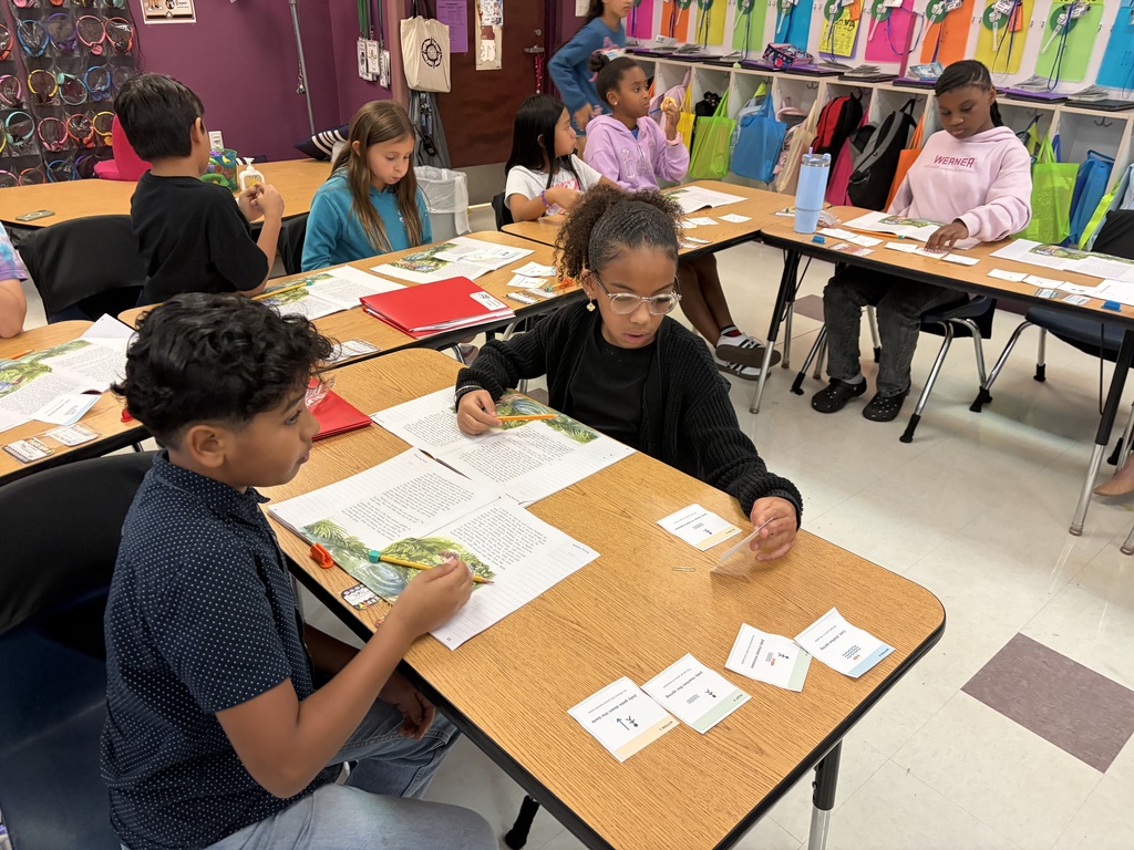 Children work on papers and books at desks in a classroom with purple walls and white tile flooring.