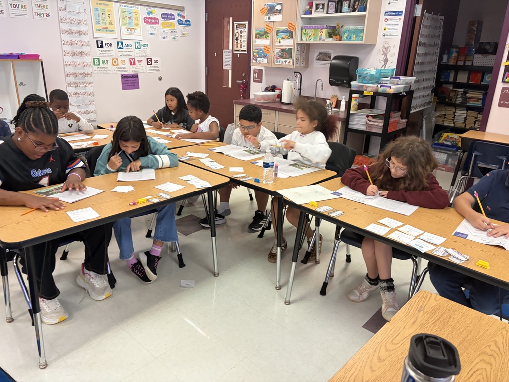 Several students in a classroom with desks and chairs, each writing on paper and using pencils.
