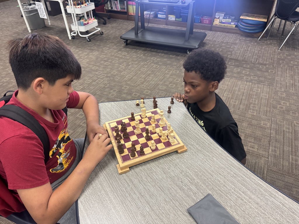 Two boys play chess on a table. One wears a red shirt, and the other a black shirt.