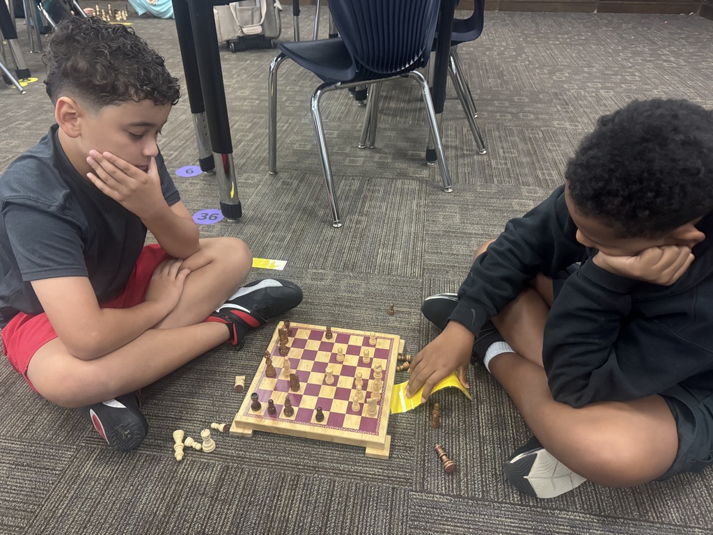 Two boys sit on the floor playing chess, one in red shorts and the other in black.