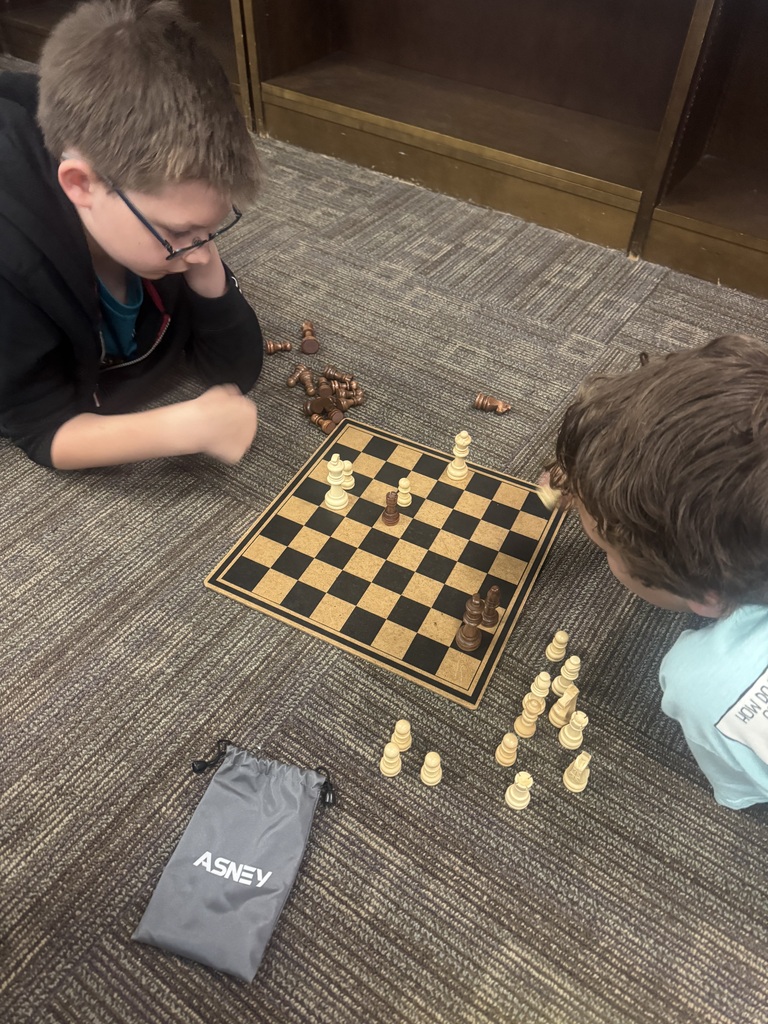 Two kids play chess on a carpeted floor. The chess set includes pieces on a board, and a bag with the word "ASHBY" is nearby.