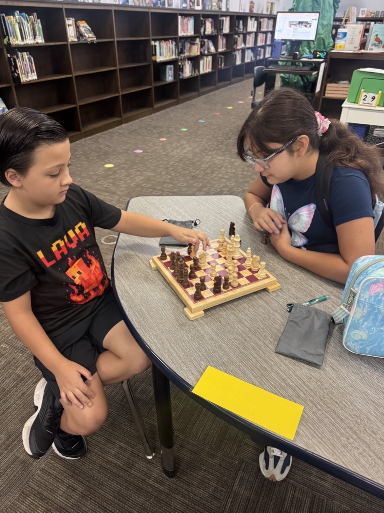Two children seated at a table in a library playing a game of chess.