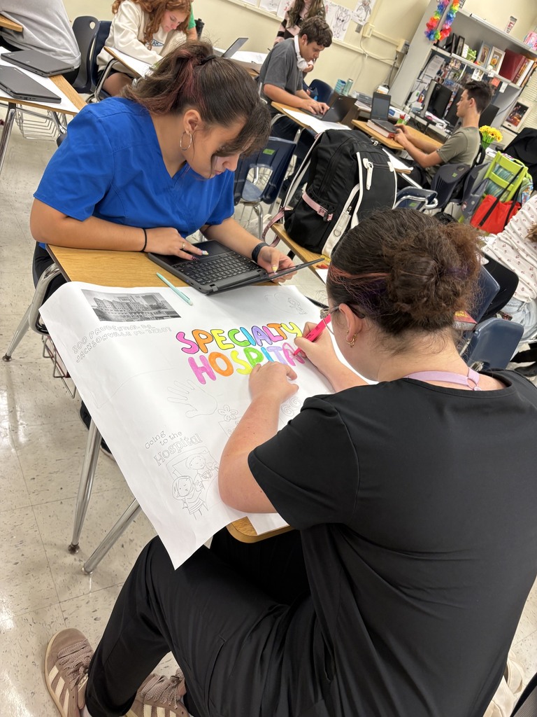 Two women sit at a desk in a classroom, each working on a large paper with colorful words.