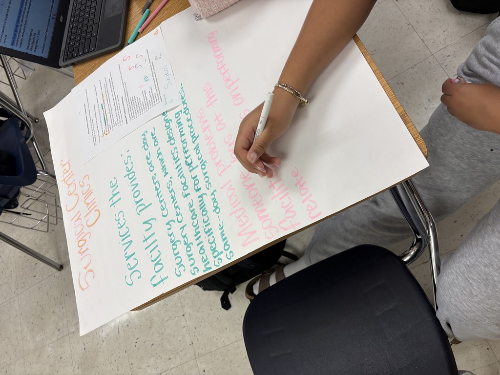 Person writing on a white board with colorful text and diagrams, using a pen. The board is on a desk, with a laptop in the background.