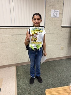A young girl with a backpack poses, holding a Safety Patrol badge with a dog. She wears jeans and sneakers.