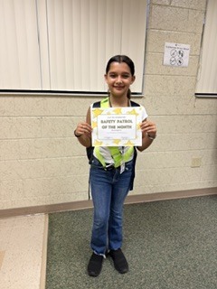 A girl poses with a certificate, wearing a safety vest and yellow safety jacket, in a room with a white wall and carpeted floor.
