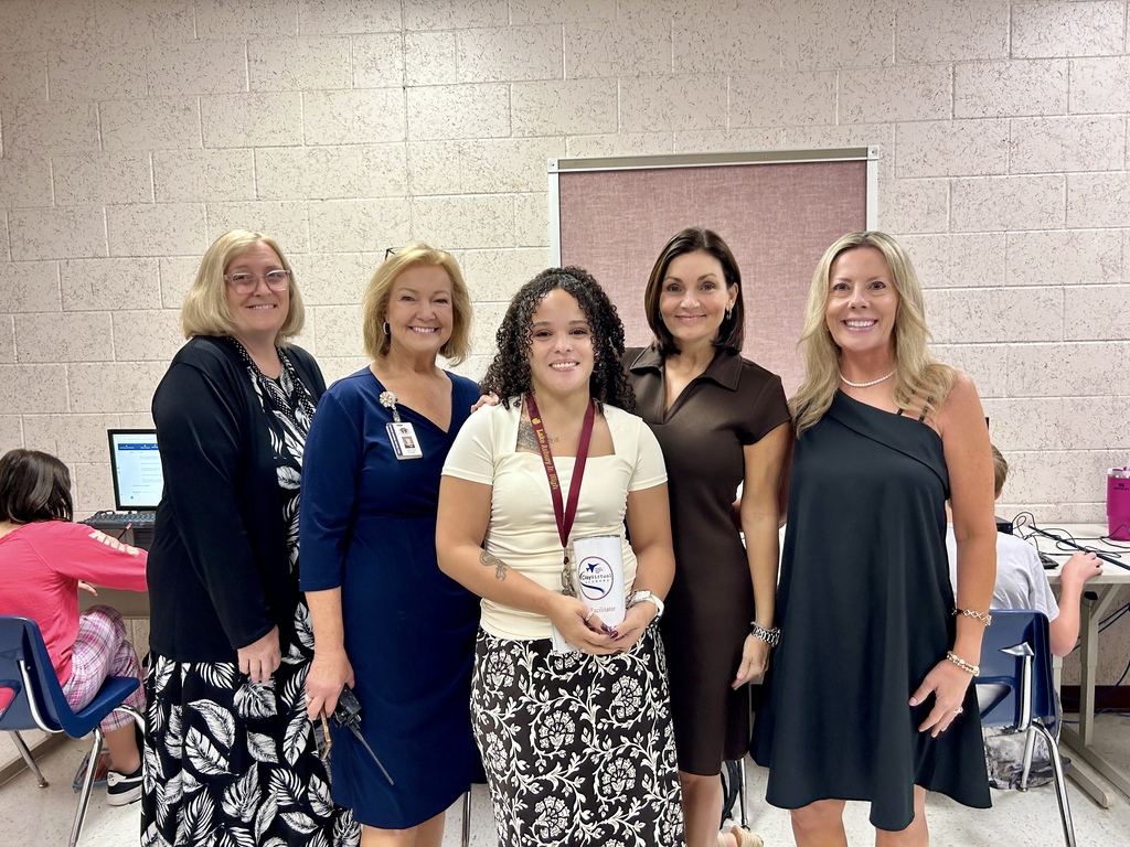 Five adult women stand together in a room with a white wall and gray tile. One woman holds an award.