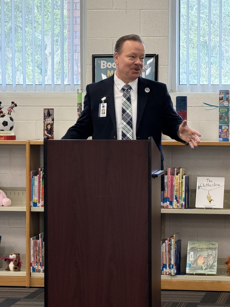 Man in suit and tie standing at podium with hands open in front of bookshelves.