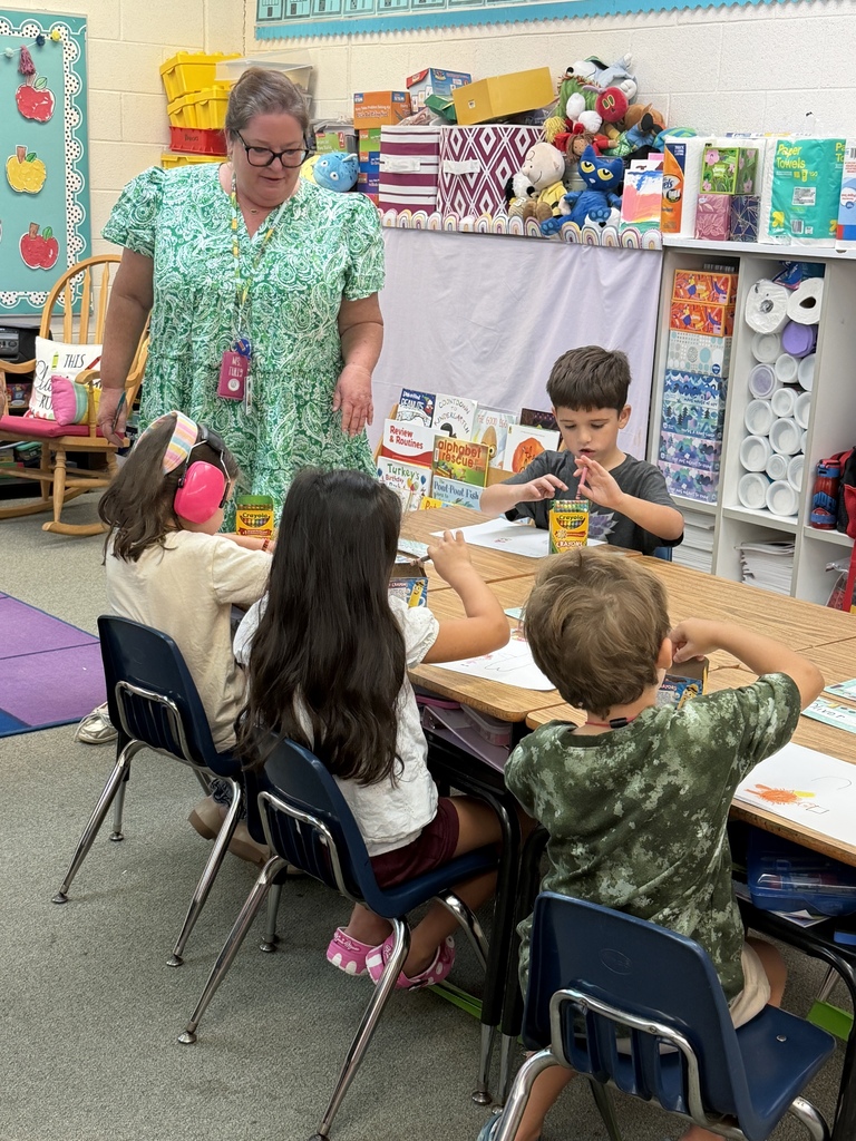 A woman with glasses and a green dress stands at a table with three children in a classroom.