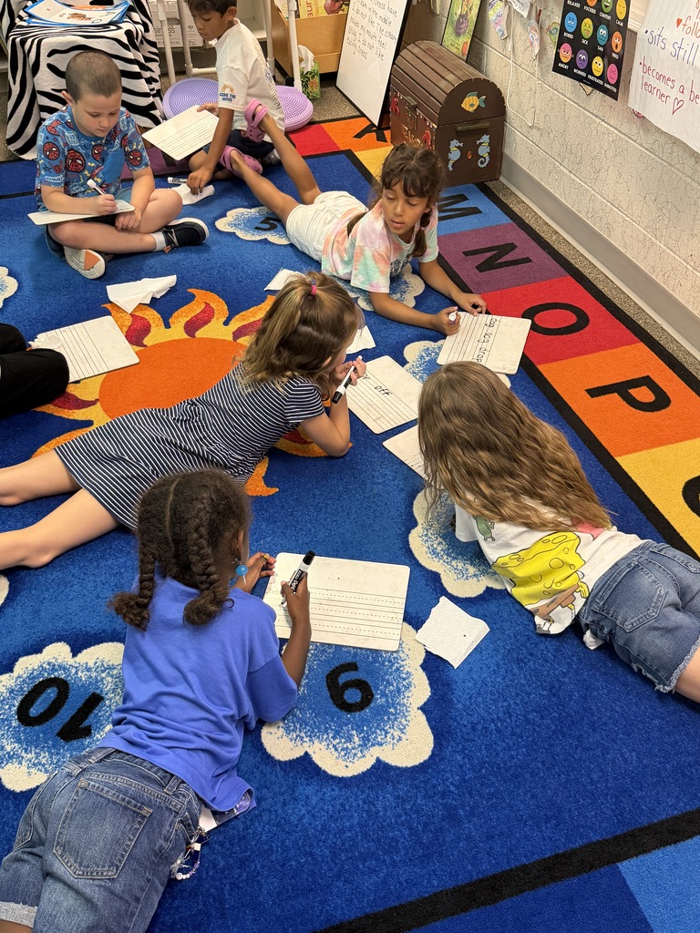 Five children sit on a blue carpet with letters and numbers. They write on paper.