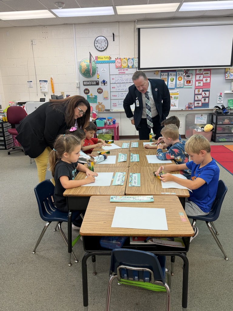 A classroom with several young children seated at desks, engaged in writing activities, and a couple of adults observing.