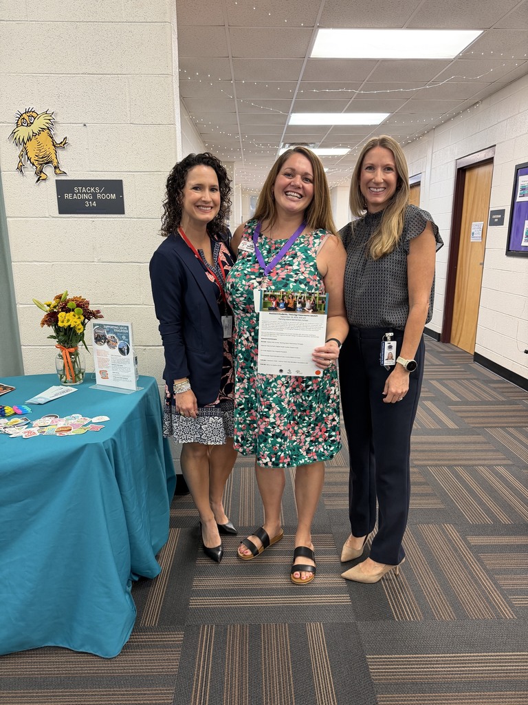 Three women pose in a hallway, one holding a certificate, with a table in the background.