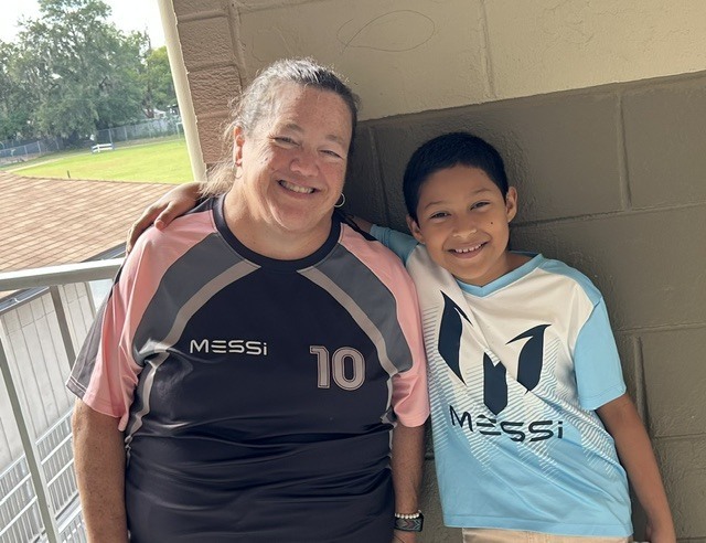 A smiling adult woman with her arm around a smiling child, both wearing Messi jerseys, standing in front of a building.