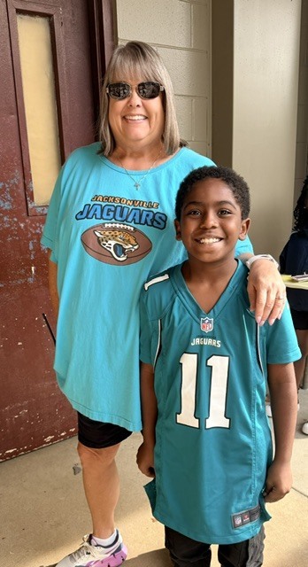 Woman and child stand close, smiling. Woman wears teal "Jacksonville Jaguars" shirt. Child wears teal "Jaguars" jersey with number 11. Behind them, door and wall.