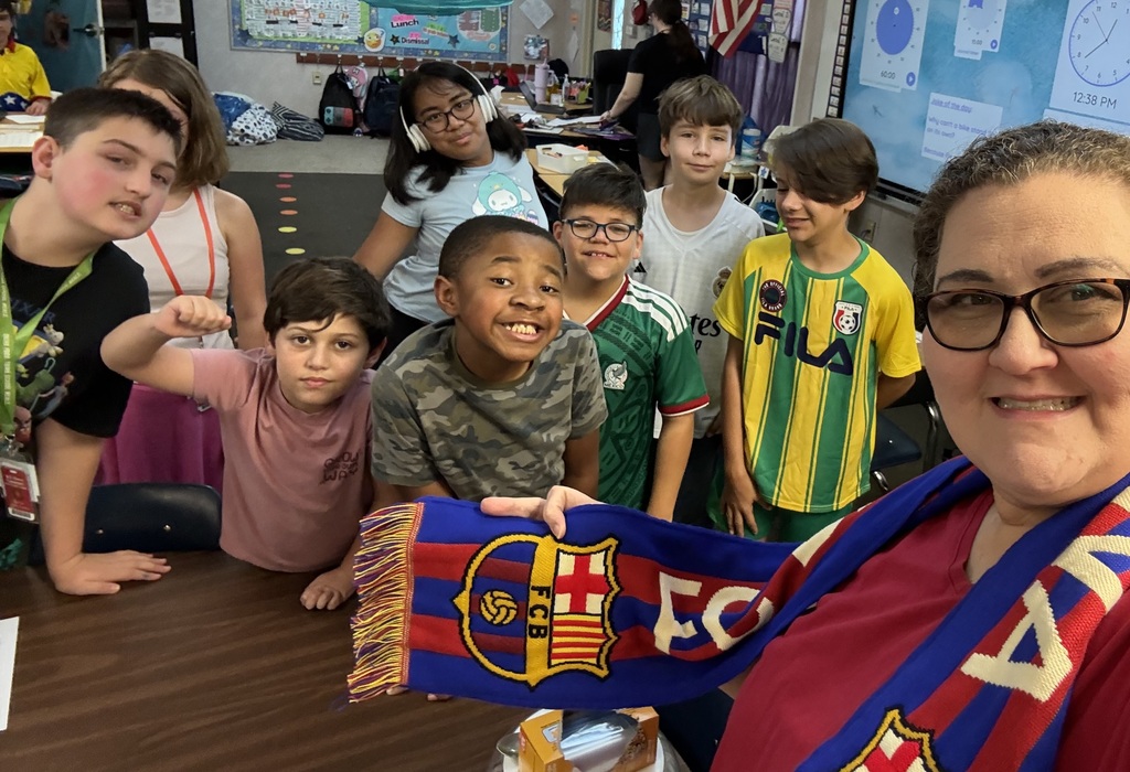 A group of children in a classroom, one holding a scarf, standing with a smiling adult.