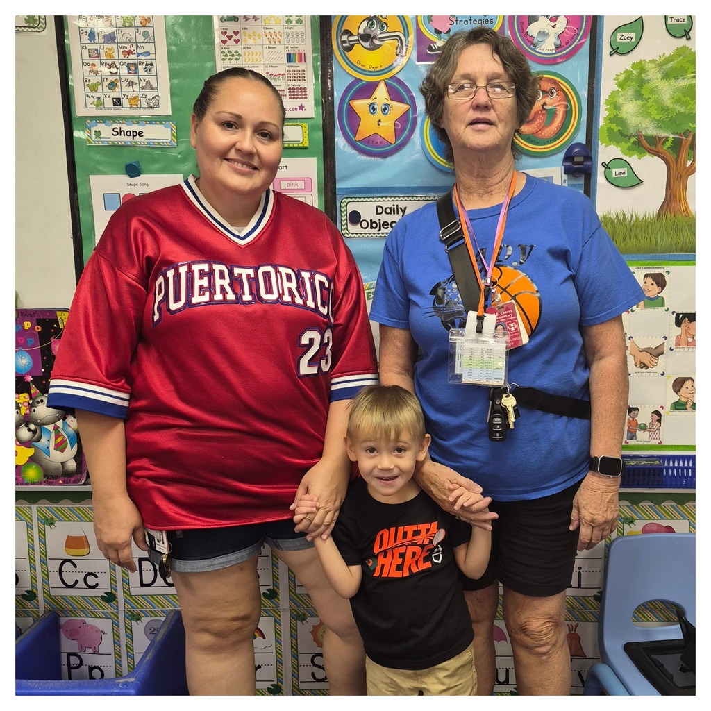 Two women and a child standing in a classroom with educational posters in the background.