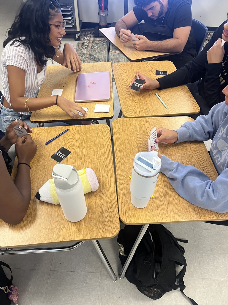 Five people sit at desks, interacting. Each person has a laptop, cards, pens, and a tumbler.