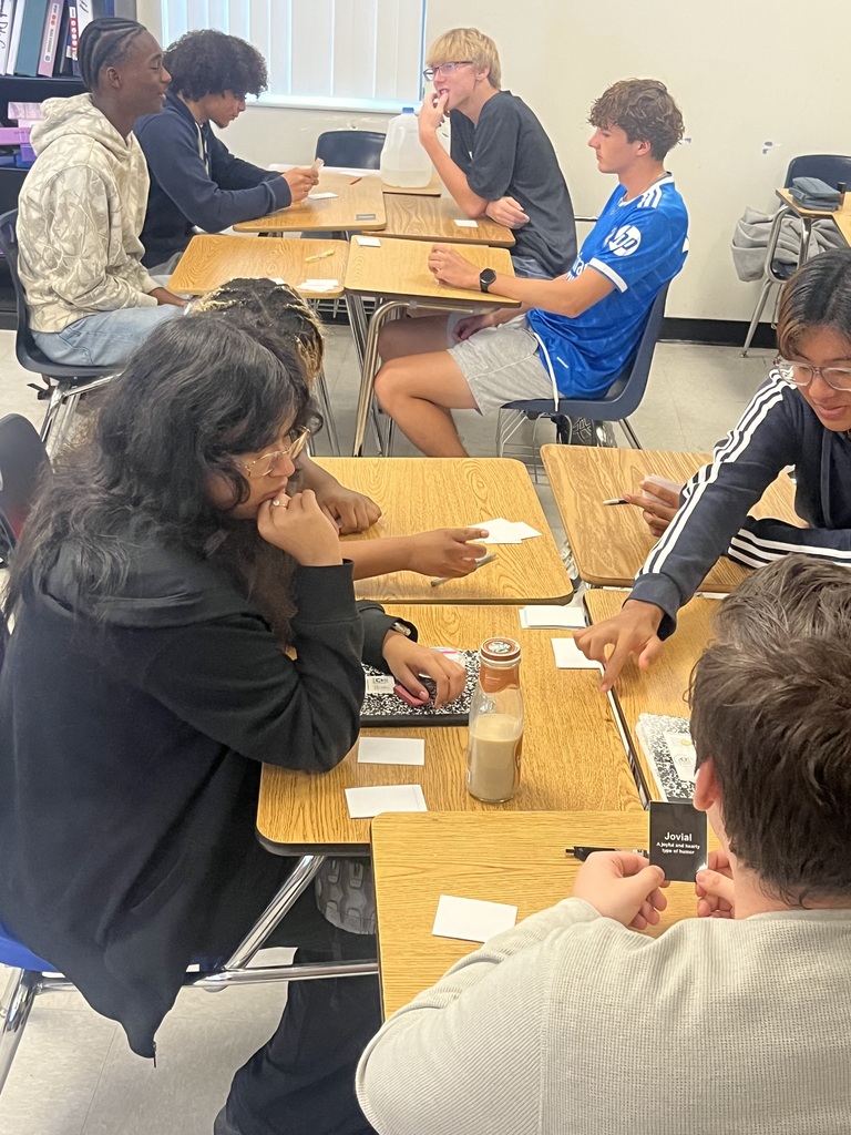 A group of students in a classroom, seated around desks, engaged in an activity using cards. The room has tables, chairs, and shelves with books.