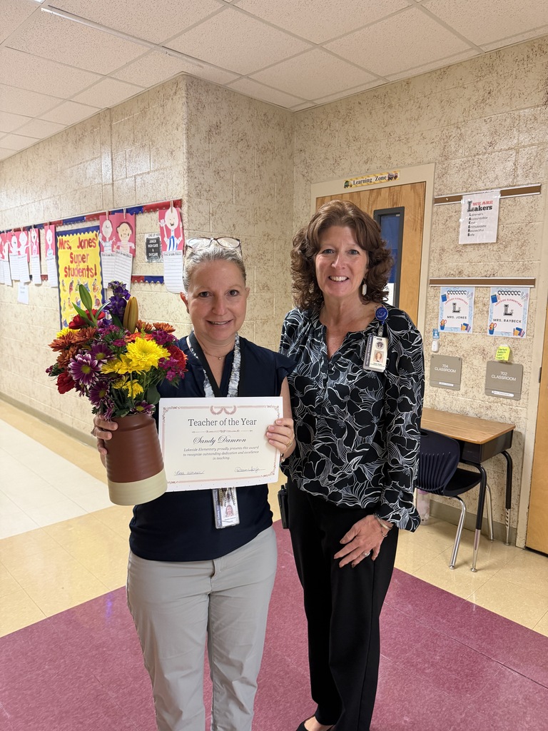 Two women stand in a hallway. One holds a bouquet and certificate. Behind them are bulletin boards with papers.
