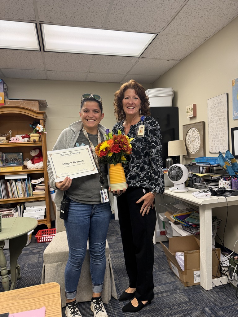 Two people standing in a room. One holds a certificate, and the other holds a flower bouquet.