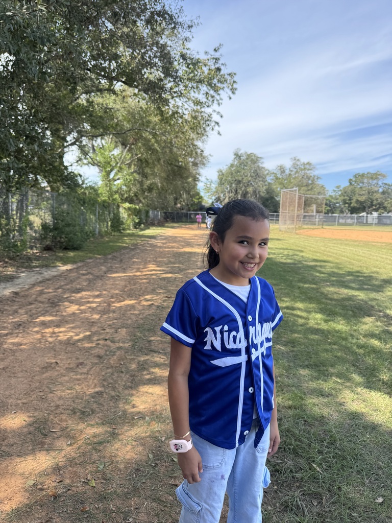 A young girl in a blue baseball jersey stands on a grassy field, near trees and a dirt path.