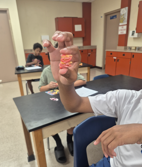 In a classroom, a person shows a hand with colorful rubber bands to another person at a desk.