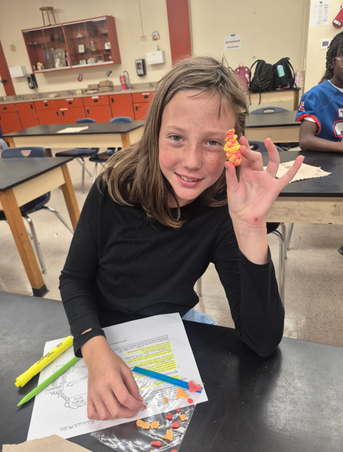 A girl in a classroom holds a multi-colored object in her hand and smiles.