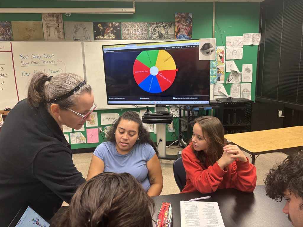 A teacher helps two students seated at a table, with a monitor displaying a colorful wheel in the background.