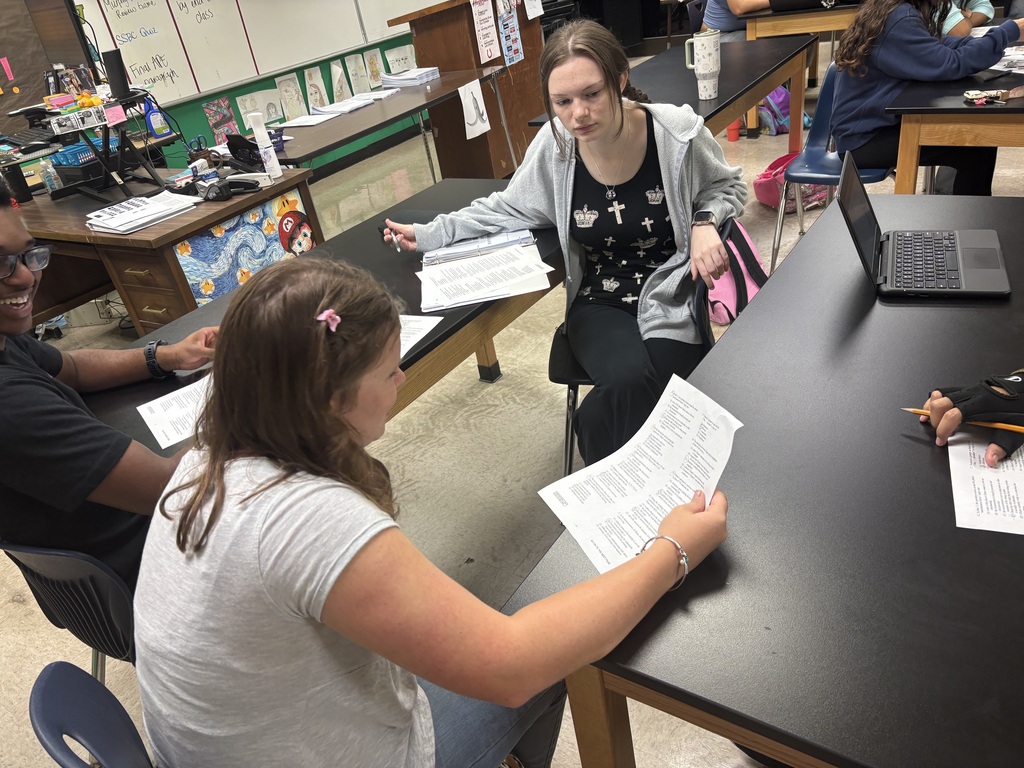 Four students in a classroom sit around tables. One woman holds papers, and another holds a laptop.