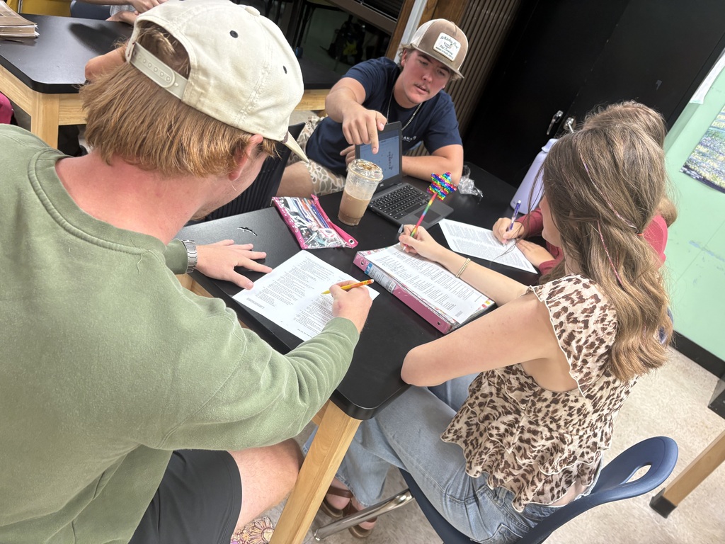 Three people sit around a table with papers, writing and using a pencil. One wears a cap, another a necklace, and the third a leopard print top.