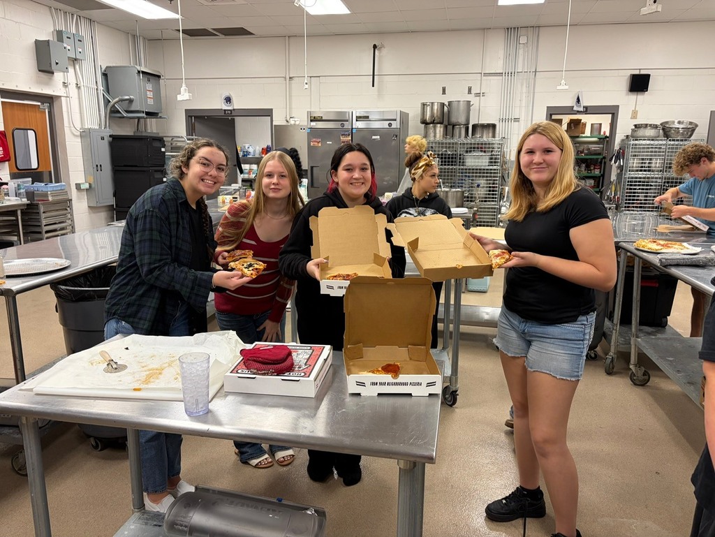 Five women in a kitchen holding food. One holds a pizza box. One holds a tray.