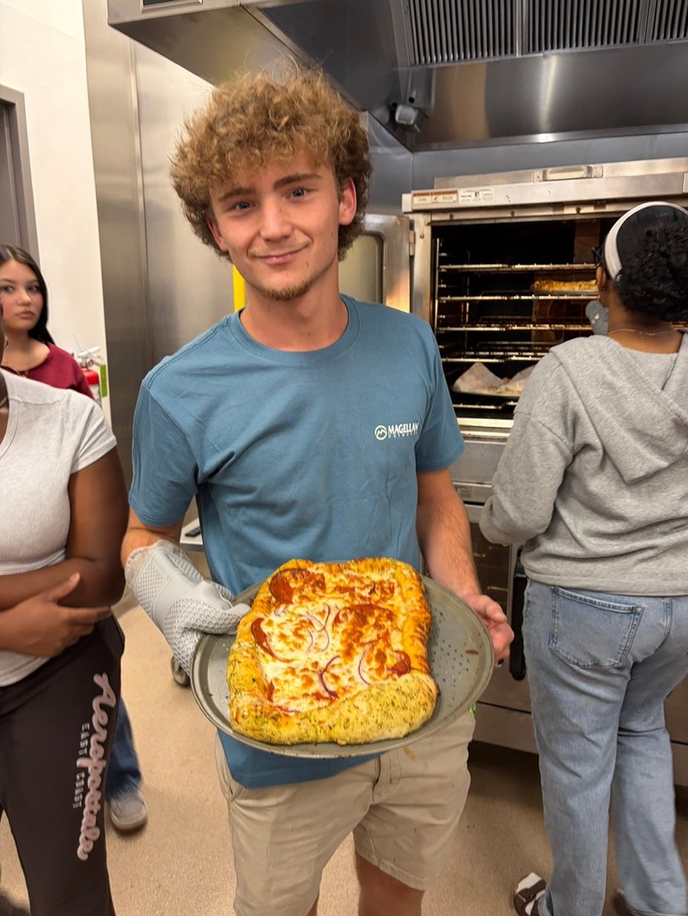 A person holds a tray with food in a kitchen. A woman stands behind him. Another person stands on the left.