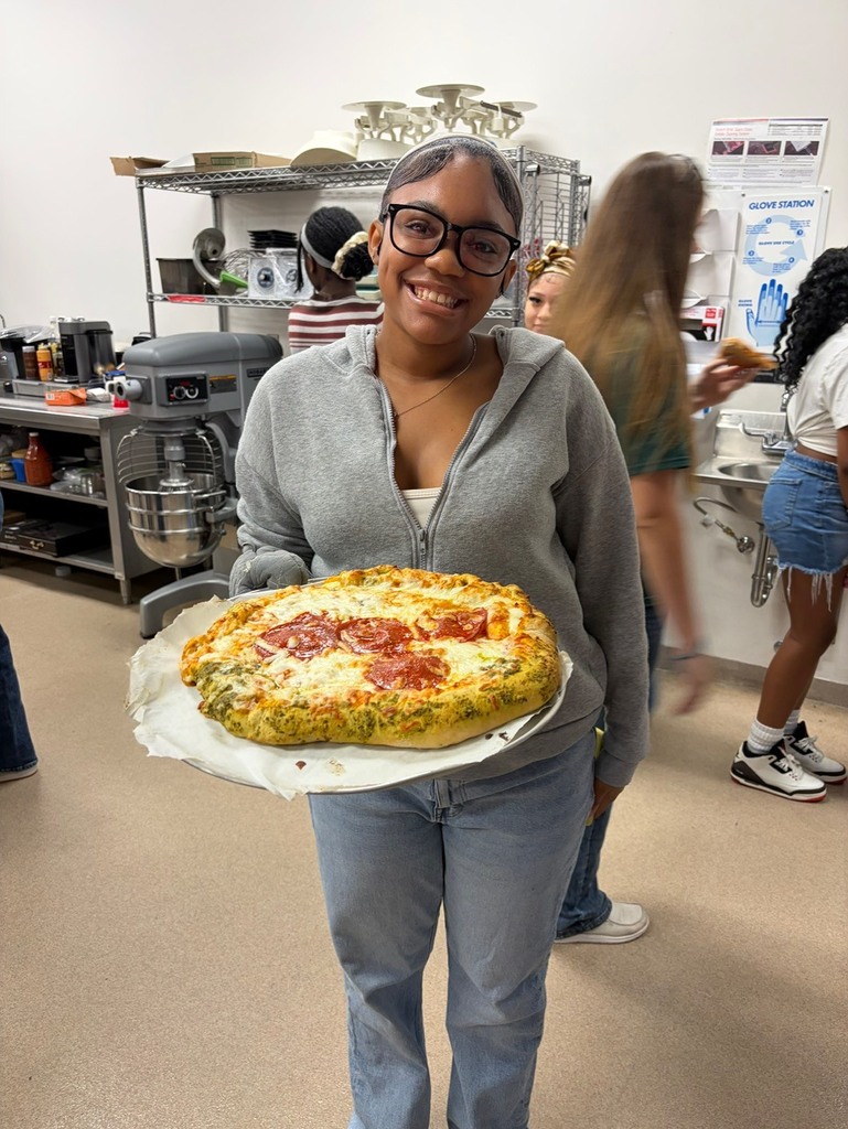 Woman holds large pizza with pepperoni and cheese toppings in a kitchen. Other people work in the background.