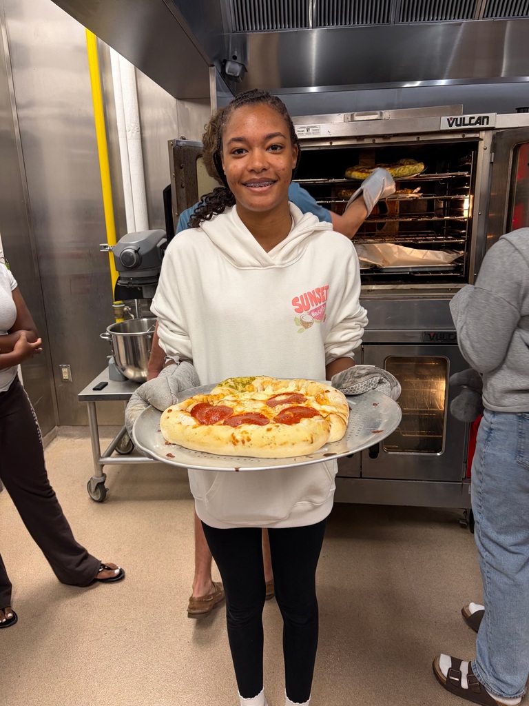 A person in a kitchen holds a pizza on a tray. An oven and another person behind them.