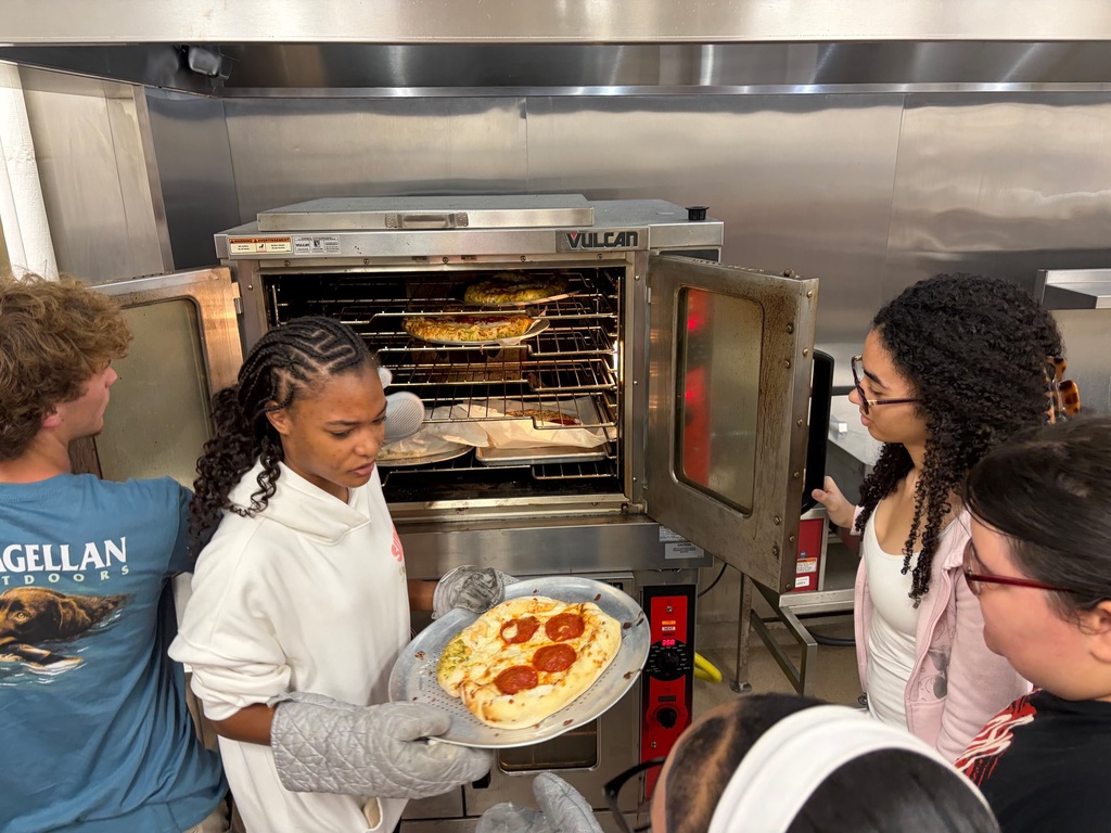 Several people in a commercial kitchen, one holds a pizza on a tray, and another has an oven door open.