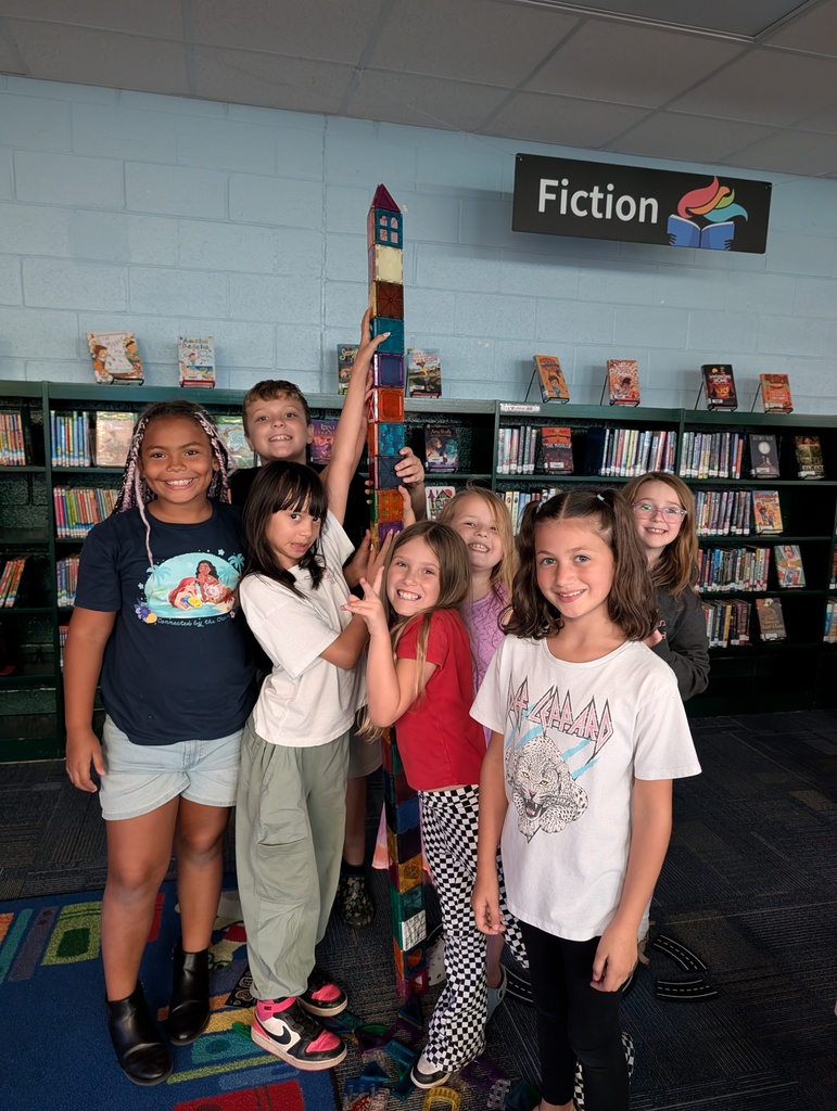 A group of children standing in a library near a bookshelf. A girl holds up a large, colorful tower.
