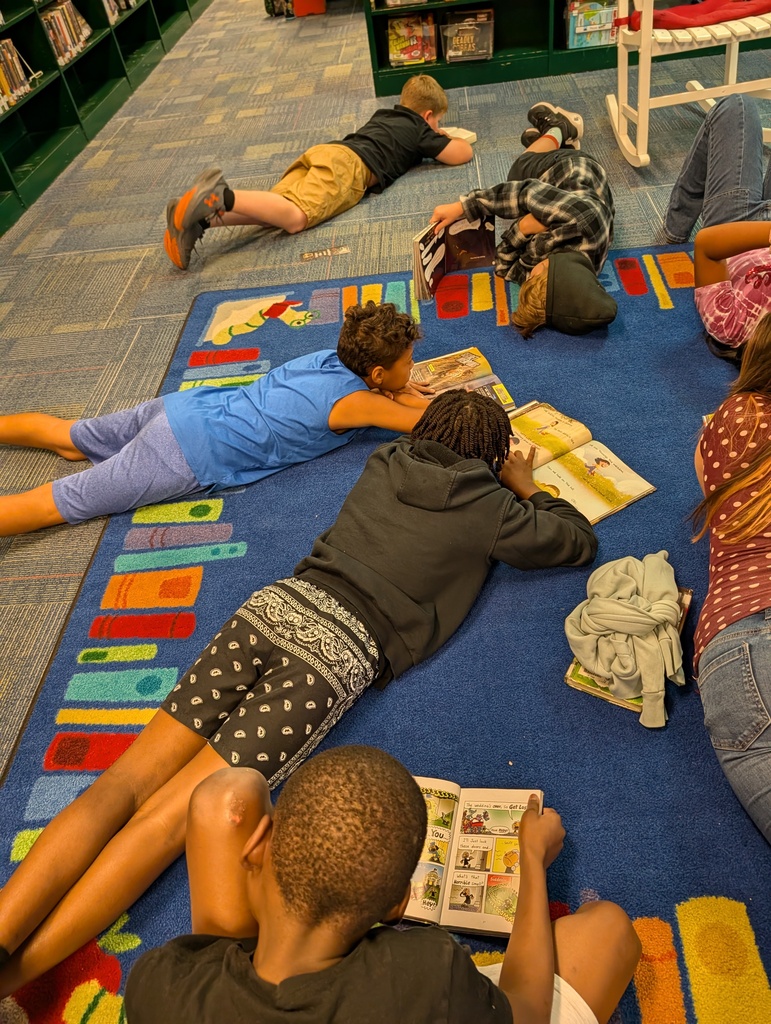 A group of children read books on a colorful carpet in a library setting. Bookshelves are in the background.