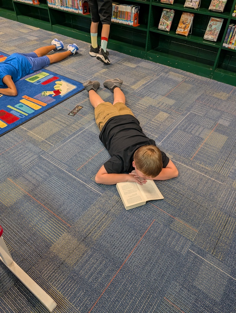 Two kids lie on the floor, reading books in a library with bookshelves behind them.