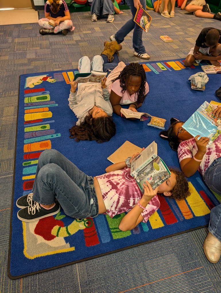 Several children lie on a multicolored floor rug, reading books. One child sits on the rug's edge.