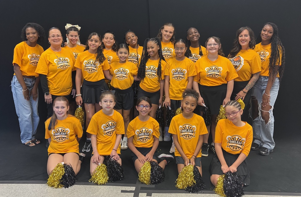 Group of cheerleaders in yellow shirts and black shorts, some holding pom-poms, posing in front of a black wall.